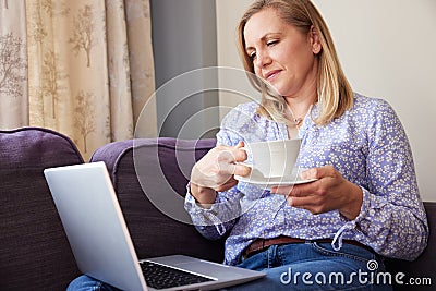 Mature Woman Sitting On Sofa Look At Laptop And Drinking Hot Drink Stock Photo