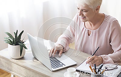 Mature working lady making notes, using laptop at home Stock Photo