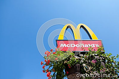 Mc Donald`s logo with its iconic M with a blue sky background and some flowers at its bottom Editorial Stock Photo