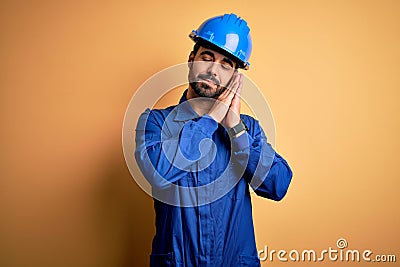 Mechanic man with beard wearing blue uniform and safety helmet over yellow background sleeping tired dreaming and posing with Stock Photo