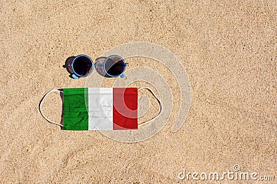A medical mask in the color of the Italian flag lies on the sandy beach next to the glasses. Stock Photo