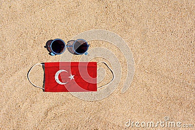 A medical mask in the color of the Turkish flag lies on the sandy beach next to the glasses. Stock Photo