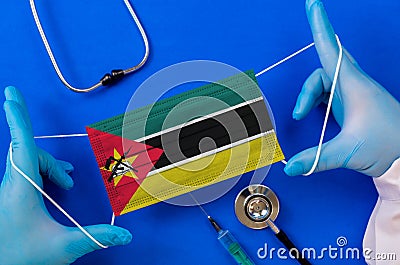 Medical mask with the flag of Mozambique in the hands of a doctor and stethoscope, flat lay. Stock Photo