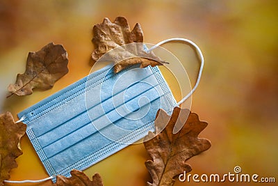 A medical protective mask and yellow dry oak leaves lie on a bright autumn background. Stock Photo