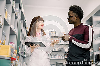 Medicine, healthcare and people concept - young woman apothecary with drug and African male customer at pharmacy Stock Photo