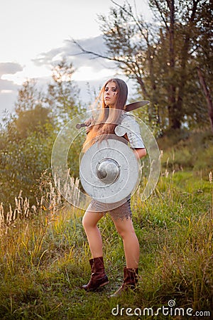 A medieval female warrior dressed in chain mail with a sword and shield in her hands poses against the background of a forest. Stock Photo