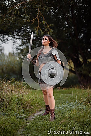 A medieval female warrior dressed in chain mail with a sword and shield in her hands poses against the background of a forest. Stock Photo