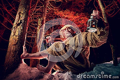 Medieval monk in canvas sackcloth robe with lattern resting on snow in dark forest and red light on winter night. Fantasy or fairy Stock Photo