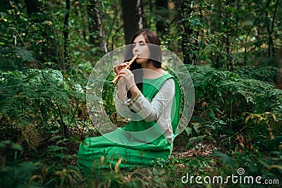 A medieval woman in a green dress plays on a wooden flute sitting in fern bushes. A girl in a gloomy forest plays music on a handm Stock Photo