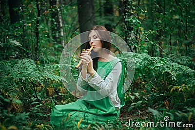 A medieval woman in a green dress plays on a wooden flute sitting in fern bushes. A girl in a gloomy forest plays music on a handm Stock Photo