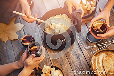 Meeting of friends, girl is cooking Swiss fondue assorted cheeses dinner on fire, hands holding mulled wine background Stock Photo