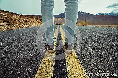 Men`s feet standing on asphalt desert road with yellow marking lines. Man wearing sneakers and jeans. Stock Photo