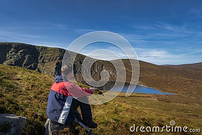 Men sitting on a rock and drinking coffee from thermos. Scenic view on Lough Ouler and Tonelagee Mountain, Ireland Stock Photo