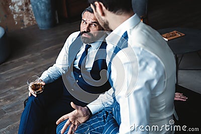 Relaxing. Two handsome men in suits holding glasses and looking at each other while resting indoors. Stock Photo