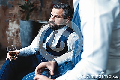 Relaxing. Two handsome men in suits holding glasses and looking at each other while resting indoors. Stock Photo