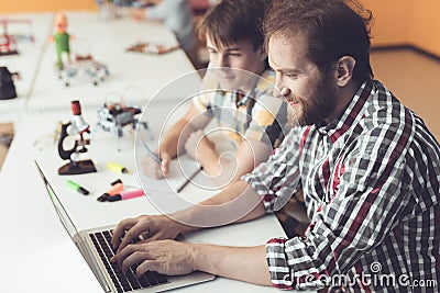 A man and a teenager are sitting at a table in front of a laptop. Nearby is a robot. They program it Stock Photo