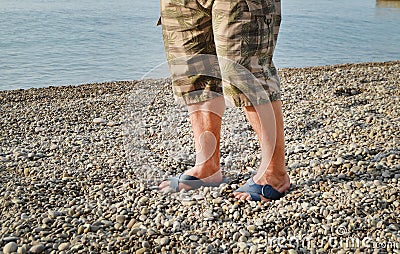 Men& x27;s feet in flip-flops and shorts, a man standing on the beach on a pebble beach, sunrise in the morning Stock Photo