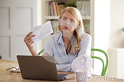 Menopausal Mature Woman Having Hot Flush At Home Cooling Herself With Fan Connected To Laptop Stock Photo