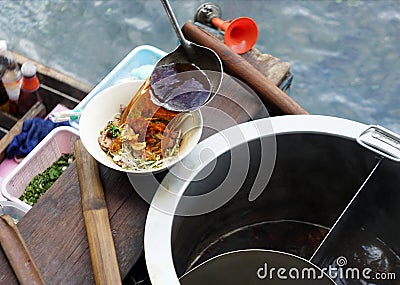 Merchant cook pouring herbal heat soup into white bowl of noodle thai style on floating wooden boat. Stock Photo