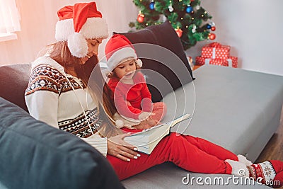 Merry Christamas and Happy New Year. Young woman with red hat sit on sofa with daughter. She holds book on her lap Stock Photo