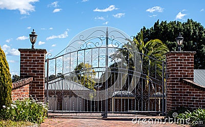 Metal driveway security entrance gates set in brick fence with residential garden in background against blue sky Stock Photo