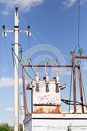 A metal rusty white elecrical transformer with wires going towards it agaisnt a blue clear sky and an electrical pole Stock Photo