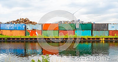 Metal scrap dump, with rusty metal parts on colorful, old truck containers, near water canal, river. Wind power windmills on backg Stock Photo