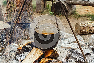Metal cauldron with meal inside hanging on tripod over firewood, preparing for boiling on bonfire in village site. Stock Photo