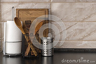 Metallic cups and kithcne utensils in it,white paper towels and wooden board on the black table against marble wall.Empty space Stock Photo