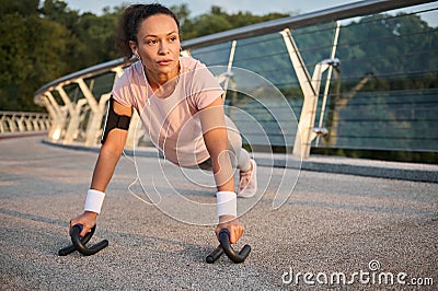 Mid adult curly haired sportswoman in pink t-shirt and gray leggins with smartphone holder and wristbands, doing push-ups outdoors Stock Photo