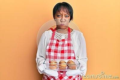 Middle age african american woman wearing baker apron holding muffins puffing cheeks with funny face Stock Photo