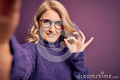 Middle age beautiful blonde woman wearing sweater and glasses make selfie by the camera doing ok sign with fingers, excellent Stock Photo