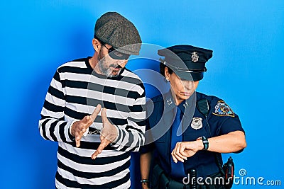 Middle age couple of hispanic woman and man wearing thief and police uniform checking the time on wrist watch, relaxed and Stock Photo