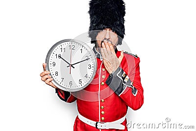 Middle age handsome wales guard man wearing traditional uniform holding big clock covering mouth with hand, shocked and afraid for Stock Photo