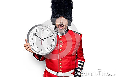 Middle age handsome wales guard man wearing traditional uniform holding big clock scared and amazed with open mouth for surprise, Stock Photo