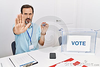 Middle age man with beard sitting by ballot holding i vote badge doing stop sing with palm of the hand Stock Photo