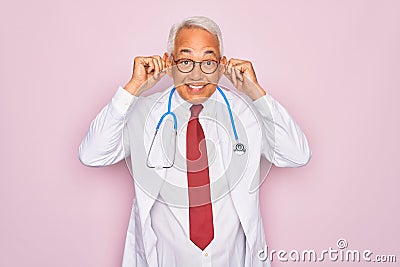 Middle age senior grey-haired doctor man wearing stethoscope and professional medical coat Smiling pulling ears with fingers, Stock Photo
