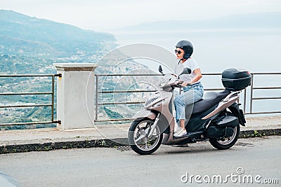 Middle-age woman in helmet and sunglasses on motor scooter on the Sicilian old town streets in the Forza d`Agro with Sant`Alessi Stock Photo