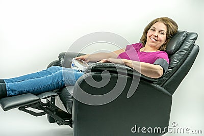 Middle-age woman leaning back in black leather recliner armchair. Checking blood pressure using portable blood pressure machine. Stock Photo