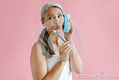 Middle aged Asian lady with bath towel holds toothbrush using sponge as phone in studio Stock Photo