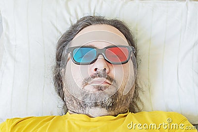 A middle-aged bearded man lies on a pillow in the early morning in colored 3D glasses Stock Photo