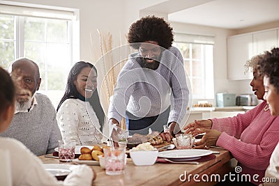 Middle aged black man bringing roast meat to the table for the Sunday family dinner with his partner, kids and their grandparents, Stock Photo