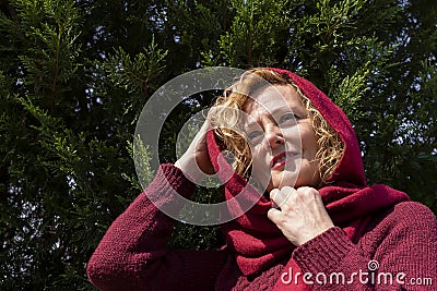 A middle-aged blonde woman plays the role of little red riding hood as in the famous fairy tale, against thuja trees Stock Photo