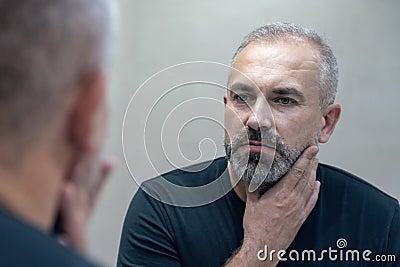 Middle-aged handsome man looking in mirror in bathroom touching his beard thinking of cutting it off Stock Photo