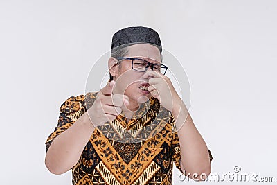 A middle aged indonesian man covers his nose while pointing at someone, complaining at their body odor. Wearing a batik shirt Stock Photo