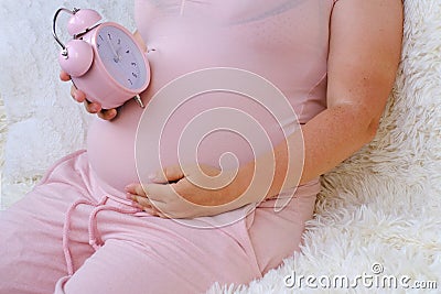 middle-aged pregnant woman holds alarm clock near her belly close-up, waiting for newborn, concept of prenatal contractions, Stock Photo