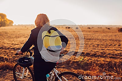 Middle-aged woman bicyclist riding in autumn field at sunset. Senior sportswoman admiring the view. Stock Photo