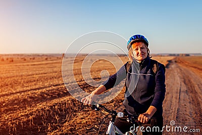 Middle-aged woman bicyclist riding in autumn field at sunset. Senior sportswoman enjoying hobby. Stock Photo