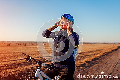 Middle-aged woman bicyclist taking on helmet in autumn field at sunset. Senior sportswoman enjoying hobby. Stock Photo