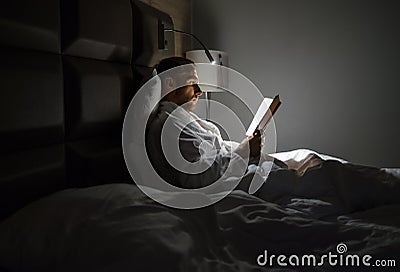 Midle-aged Caucasian man relaxing in bed reading bestseller novel paper book with bedside lamp turned on. Evening relaxation, Stock Photo